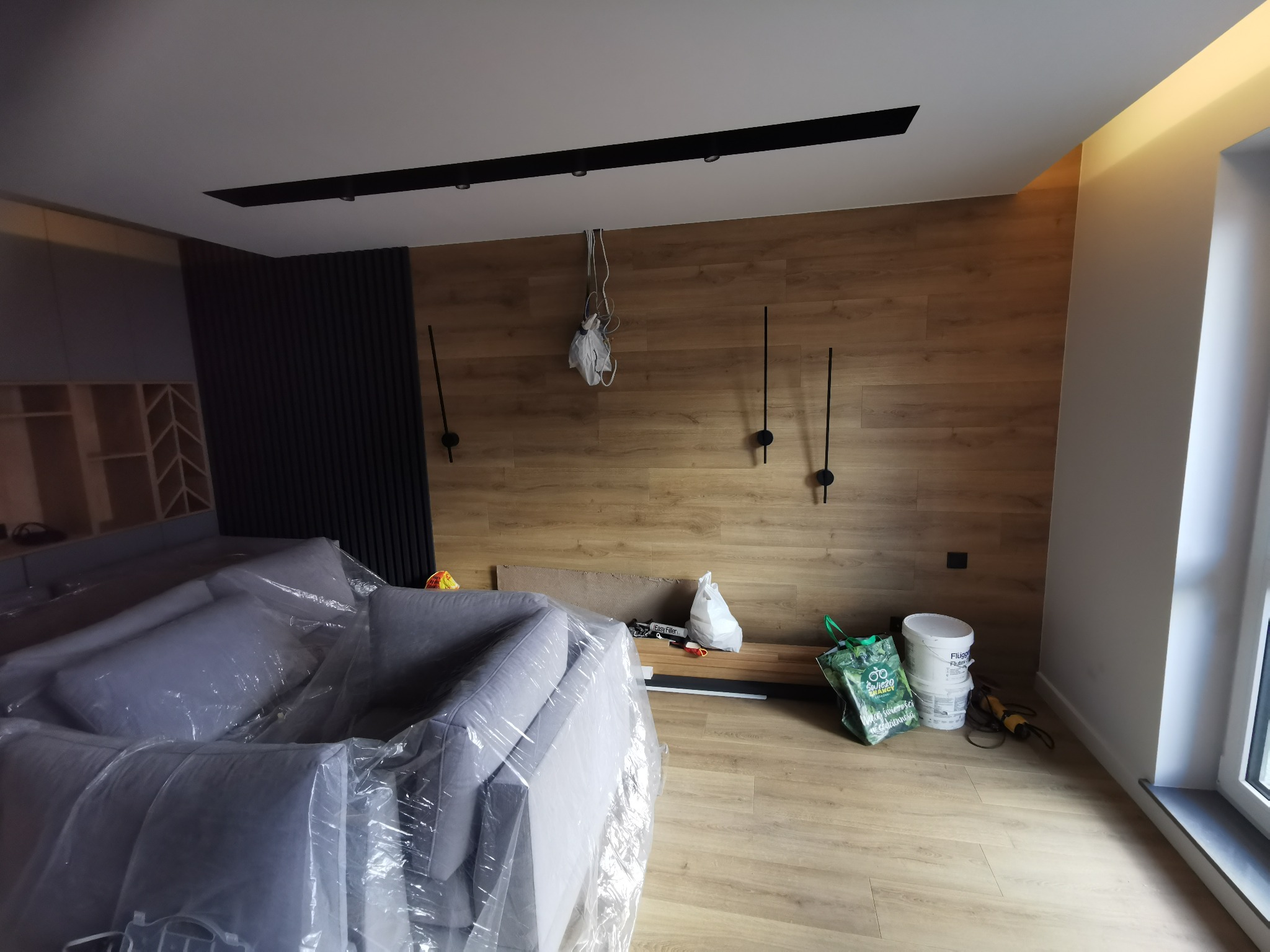 Pokój w trakcie prac wykończeniowych: zabezpieczona folią sofa, drewniana okładzina ścienna, wiszące kable, puszki z farbą i narzędzia na podłodze.