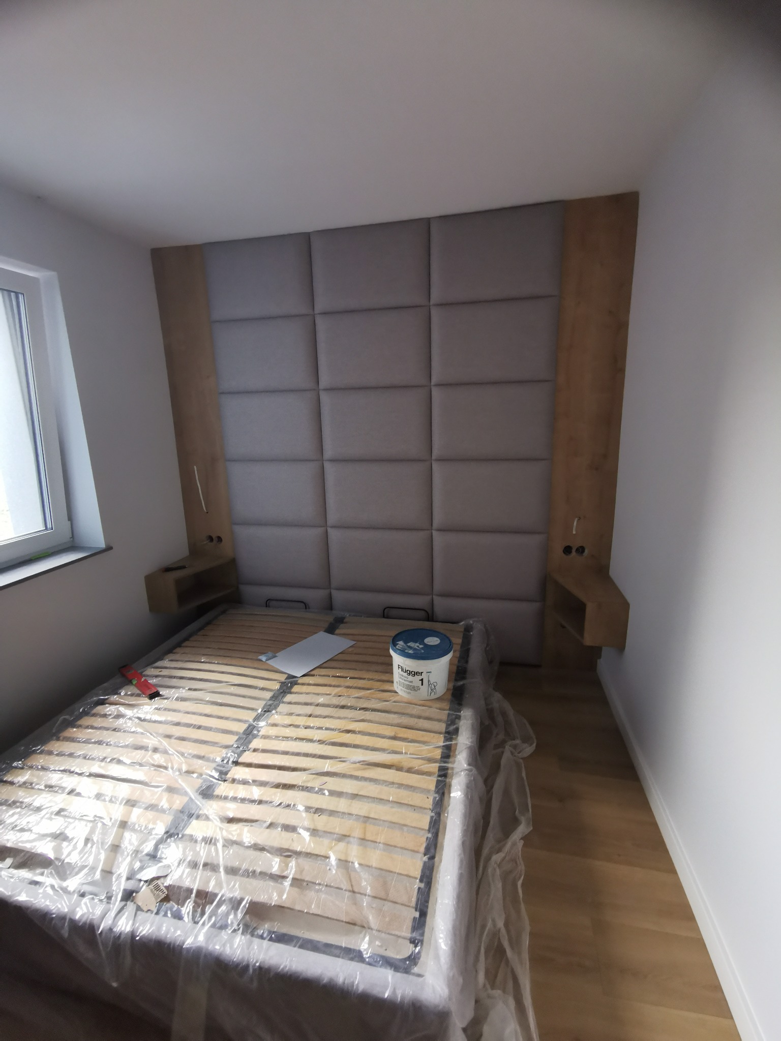 Pokój w trakcie przygotowań do malowania: łóżko przykryte folią ochronną, widoczna puszka z farbą Flugger i zagłówek z oświetleniem.