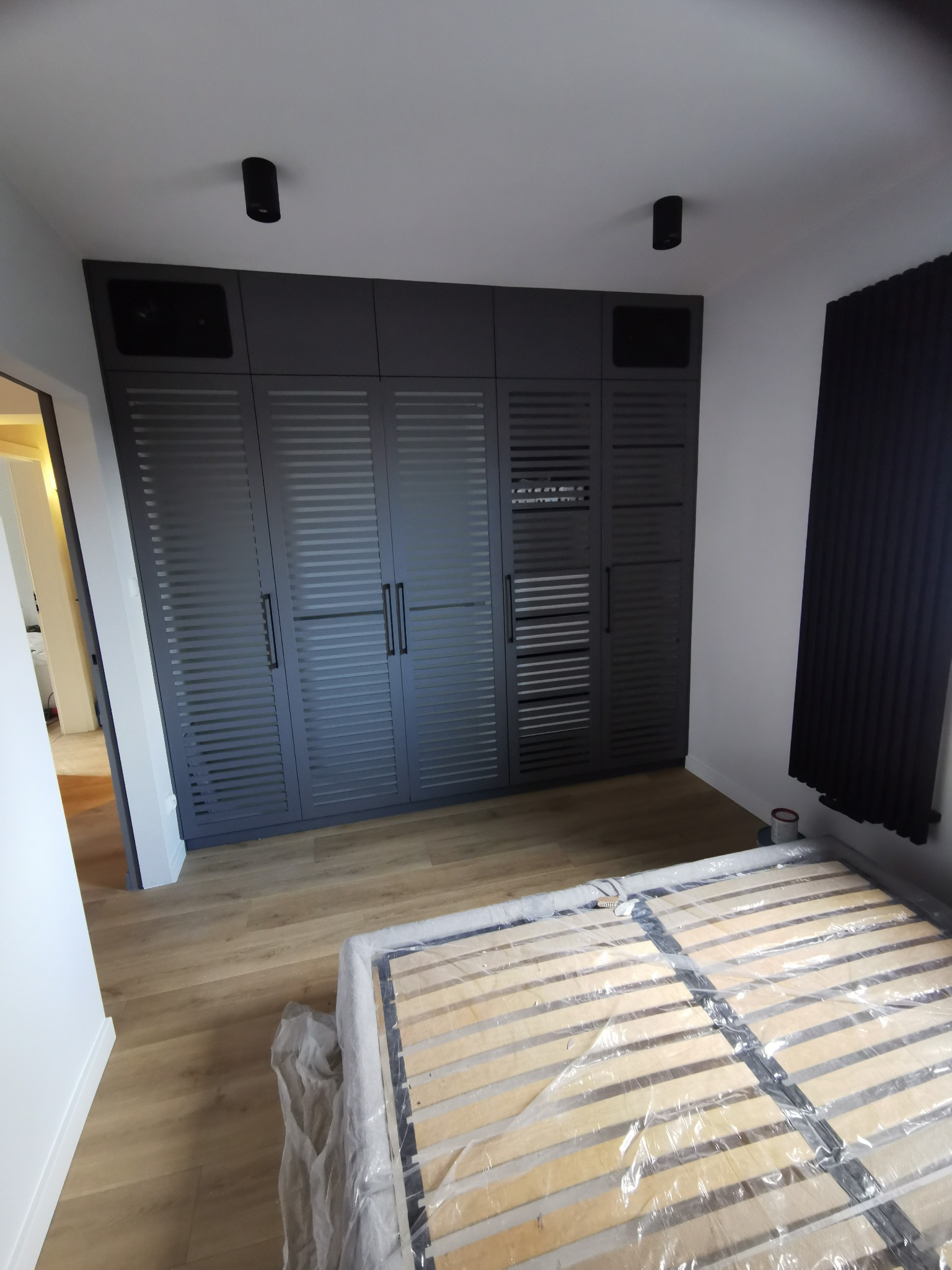 Szara zabudowa meblowa z lamelami w sypialni, widoczny stelaż łóżka zabezpieczony folią, dwa czarne reflektory sufitowe i grzejnik żeberkowy w tle.