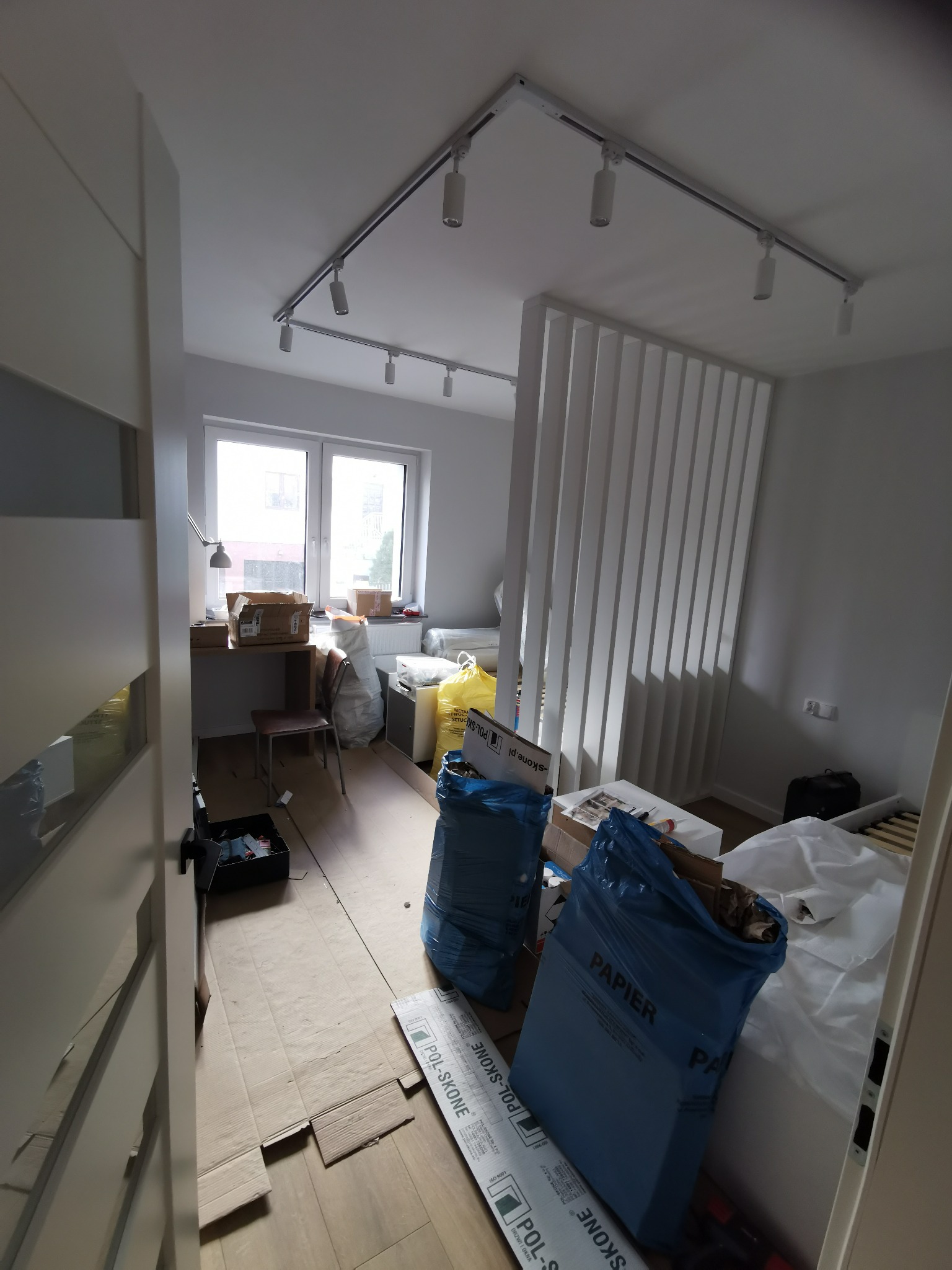 Pokój w trakcie przygotowań do prac wykończeniowych, zabezpieczony kartonami i folią, widoczne worki na śmieci, oświetlenie szynowe.