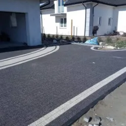 Czarna nawierzchnia z jasnymi pasami oddzielającymi, prowadząca do garażu i domu z białymi elewacjami i ciemnym dachem, widoczne niedokończone prace brukarskie.