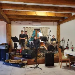 Zespół muzyczny grający na scenie podczas imprezy, widoczne saksofony w futerałach, perkusja, keyboard i gitara.