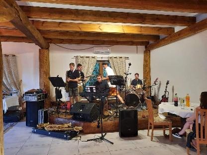 Zespół muzyczny grający na drewnianej scenie w rustykalnym wnętrzu z drewnianymi belkami stropowymi, widoczne instrumenty: saksofon, keyboard, perkusja i gitara, nagłośnienie oraz siedząca przy...