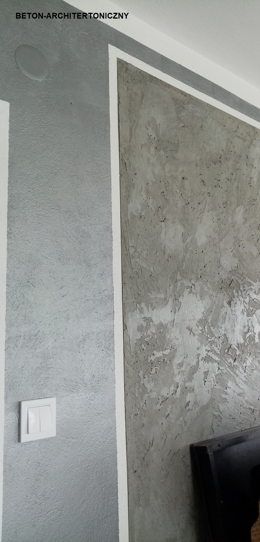 Fragment ściany z szarą, chropowatą fakturą betonu architektonicznego, oddzielony białą listwą od gładkiej, szarej ściany z włącznikiem światła. W górnym rogu widoczny głośnik sufitowy.