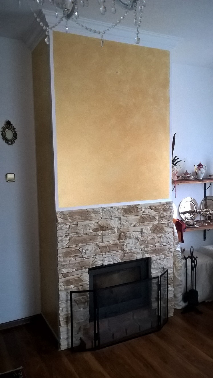 Otynkowany kominek z obudową z kamienia dekoracyjnego i czarnym ekranem ochronnym, zwieńczony złotą ścianą, oświetlony kryształowym żyrandolem.