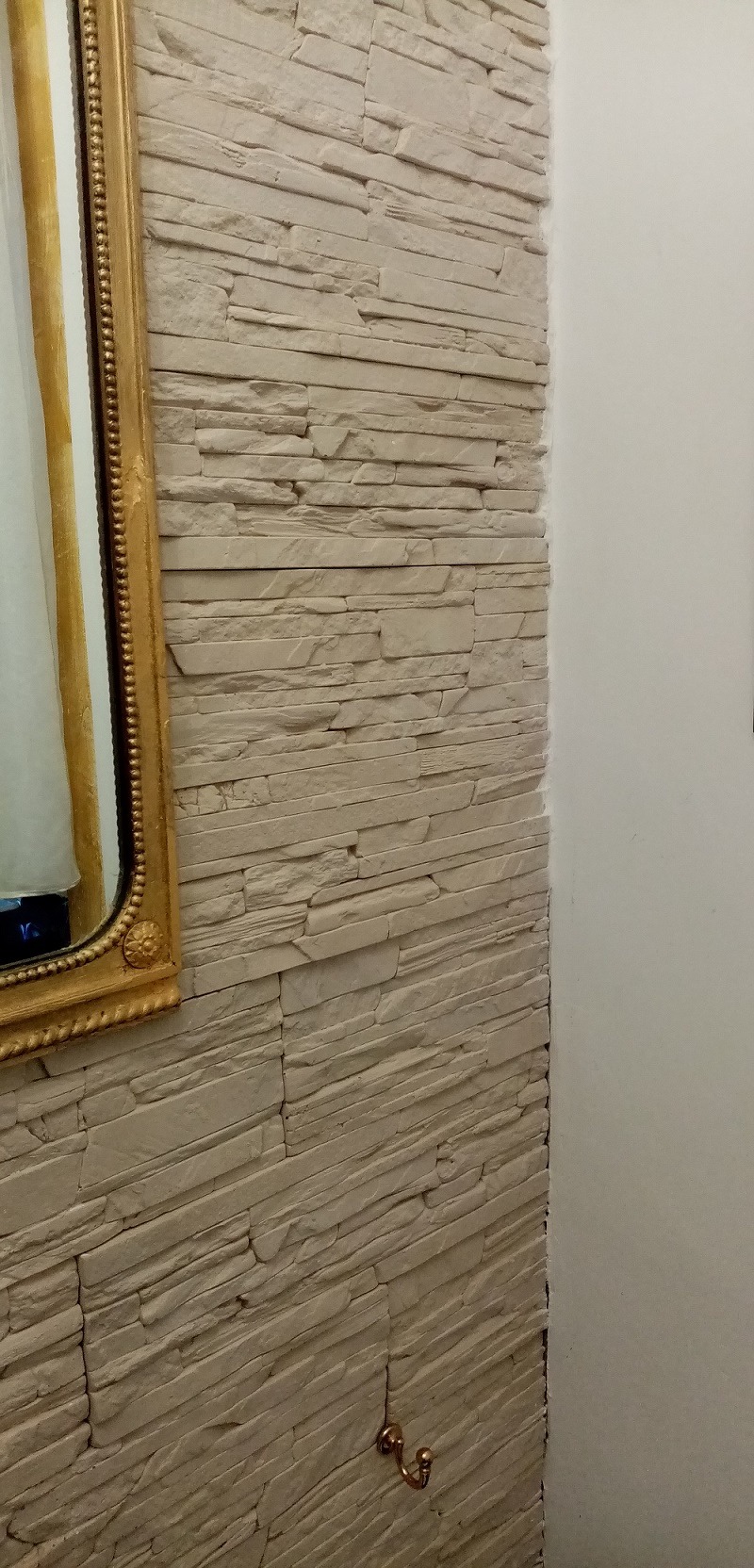 Ściana wyłożona dekoracyjnym kamieniem w odcieniach beżu, z widocznym fragmentem lustra w złotej ramie i małym, metalowym haczykiem na ścianie.