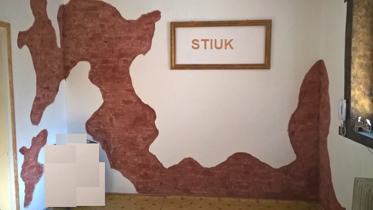 Fragment ściany z dekoracyjnym stiukiem w kolorze ceglastym, nieregularne kształty na białym tle, nad nim ramka z napisem 'STIUK', widoczny fragment pokoju z dywanem na podłodze.