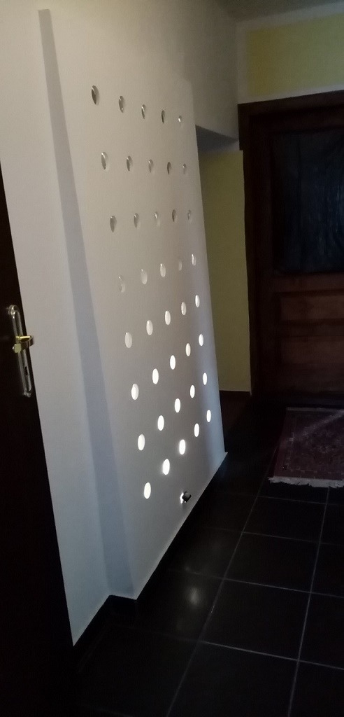 Biała, podświetlana ściana działowa z otworami w kształcie kropli, widoczna z perspektywy podłogi w korytarzu z ciemnymi płytkami.