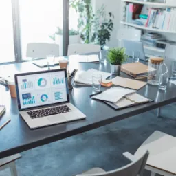 Laptop wyświetlający dashboard z wykresami na stole konferencyjnym, otoczony notatnikiem, długopisem, dokumentami i szklankami wody, sugerujący analizę danych i planowanie strategiczne.