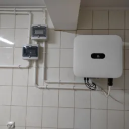 Biała skrzynka inwertera Huawei zamontowana na kafelkowanej ścianie, widoczne białe rury instalacyjne i dwie szare skrzynki zabezpieczeń elektrycznych.