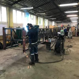 Spawacz w kombinezonie spawalniczym łączy elementy stalowe konstrukcji hali w warsztacie, widoczne iskry i dym spawalniczy, w tle maszyna spawalnicza i drugi pracownik.