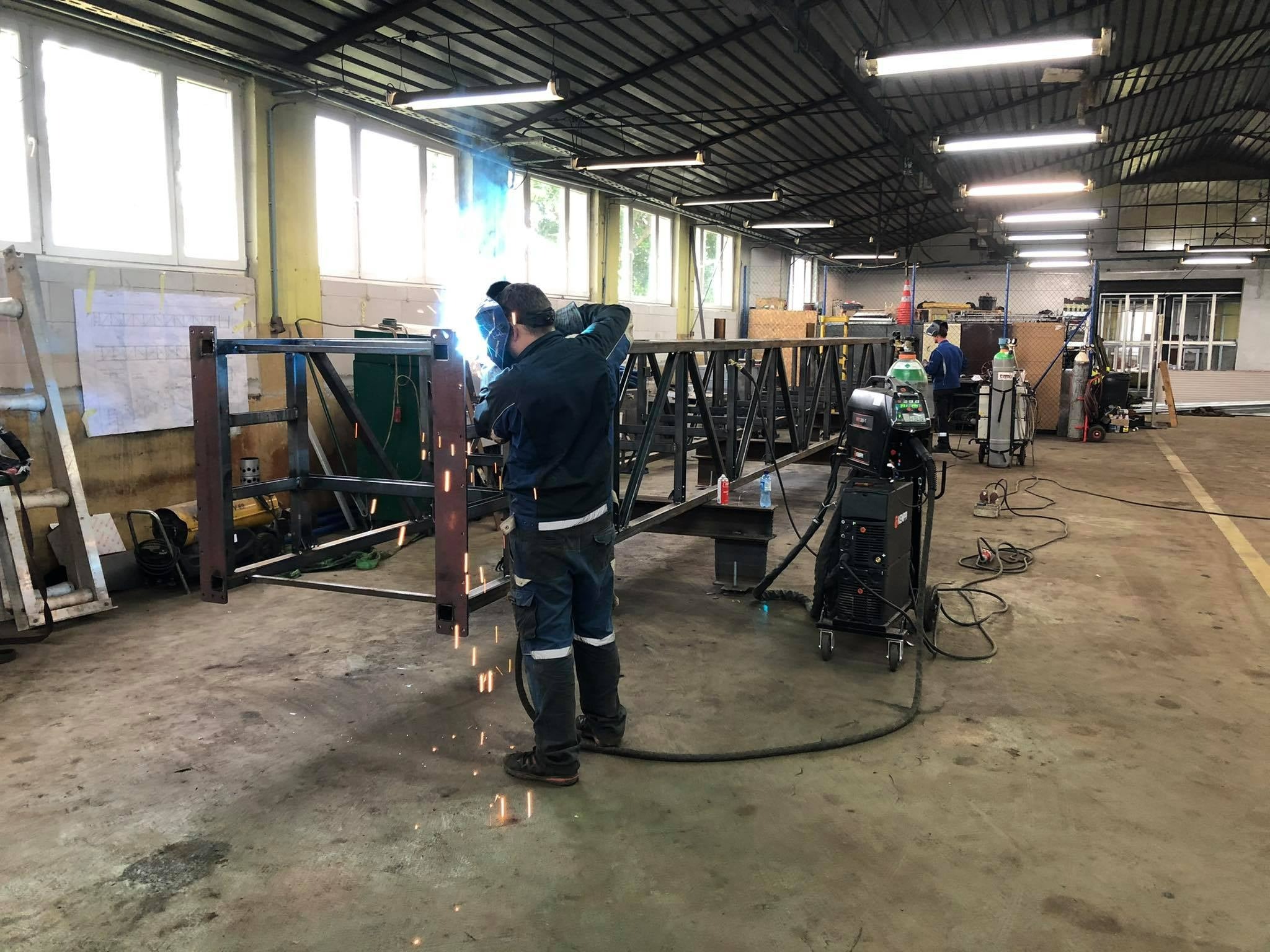 Spawacz w kombinezonie spawalniczym łączy elementy stalowe konstrukcji hali w warsztacie, widoczne iskry i dym spawalniczy, w tle maszyna spawalnicza i drugi pracownik.