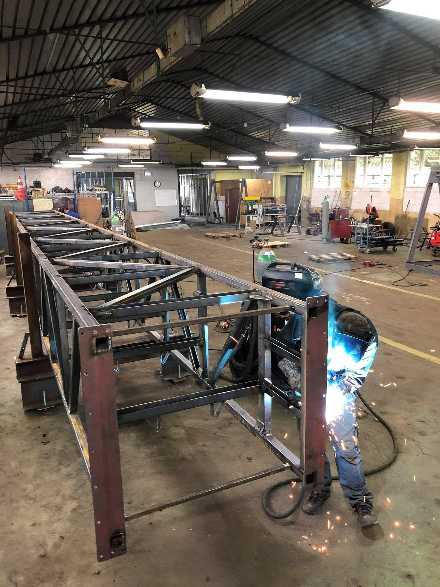 Pracownik w kombinezonie spawalniczym łączy elementy stalowej konstrukcji w hali, widoczne iskry i spawarka WFX 300-T.