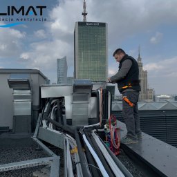 I-Klimat sp. z o.o. - Instalator w kamizelce i spodniach roboczych, z pomarańczowym narzędziem przy pasie, pracuje na dachu budynku w centrum Warszawy, przy instalacji wentylacyjnej; w tle widoczne wieżowce, w tym...