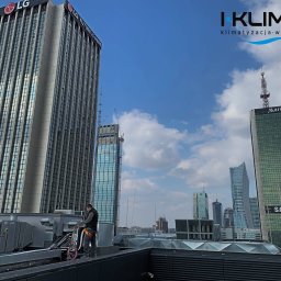I-Klimat sp. z o.o. - Pracownik serwisu w uprzęży na dachu budynku, wykonujący prace konserwacyjne przy systemie wentylacji i klimatyzacji na tle panoramy Warszawy z wieżowcami LG, Marriott i Samsung.