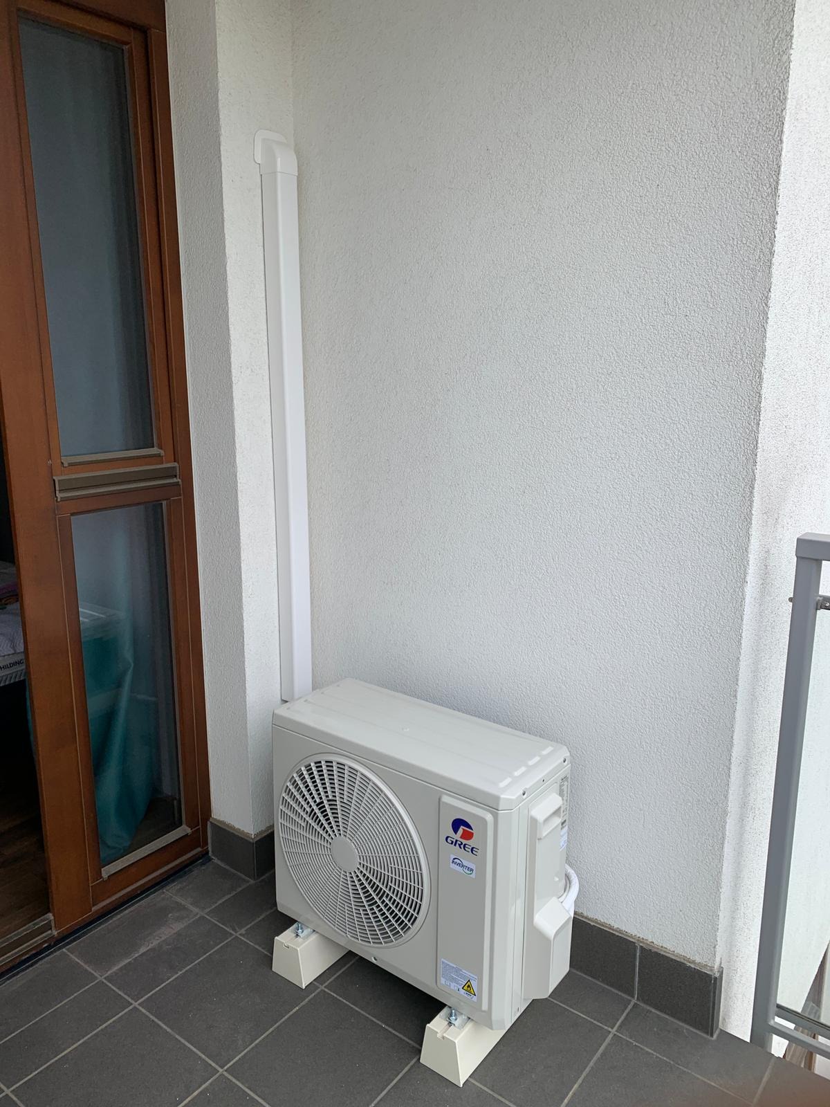 Kompaktowa, biała jednostka zewnętrzna klimatyzacji marki Gree, zamontowana na betonowym balkonie na podwyższeniu z białych bloczków, z widoczną rurą odprowadzającą skropliny przy ścianie budynku.
