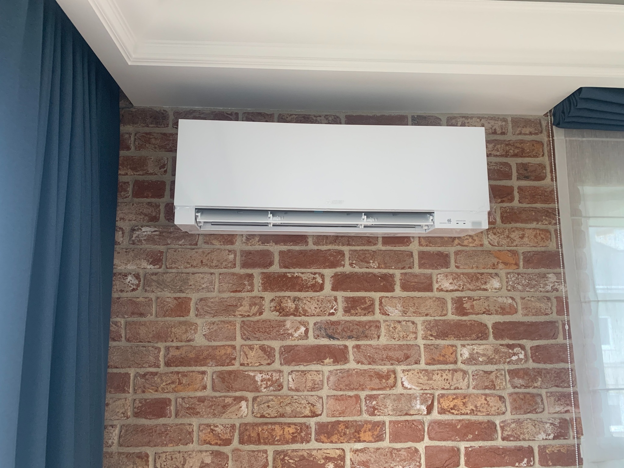 Biała jednostka klimatyzacji Mitsubishi Electric zamontowana na ścianie z czerwonej cegły w nowoczesnym wnętrzu z niebieskimi zasłonami.