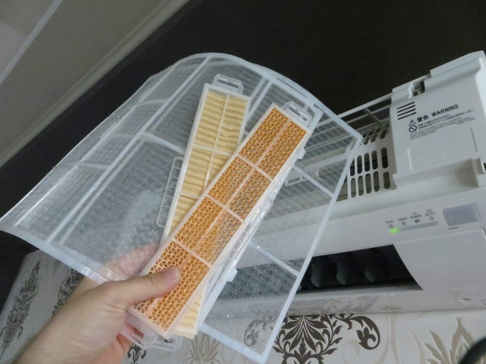 Filtr powietrza klimatyzatora, z widocznymi zabrudzeniami, wyjmowany do czyszczenia, na tle białej jednostki wewnętrznej z zieloną diodą.