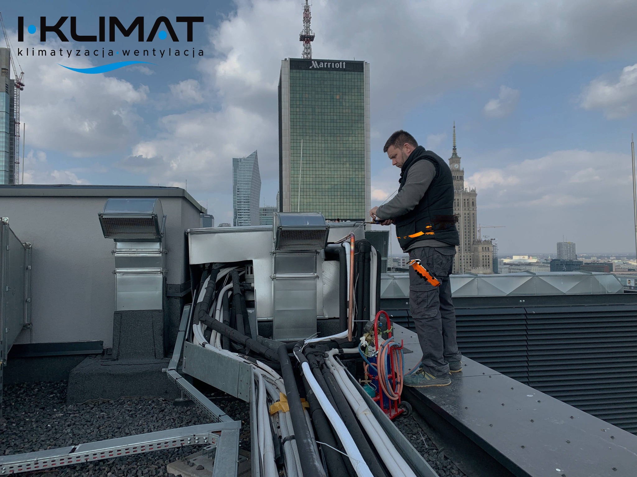 Instalator w kamizelce i spodniach roboczych, z pomarańczowym narzędziem przy pasie, pracuje na dachu budynku w centrum Warszawy, przy instalacji wentylacyjnej; w tle widoczne wieżowce, w tym...