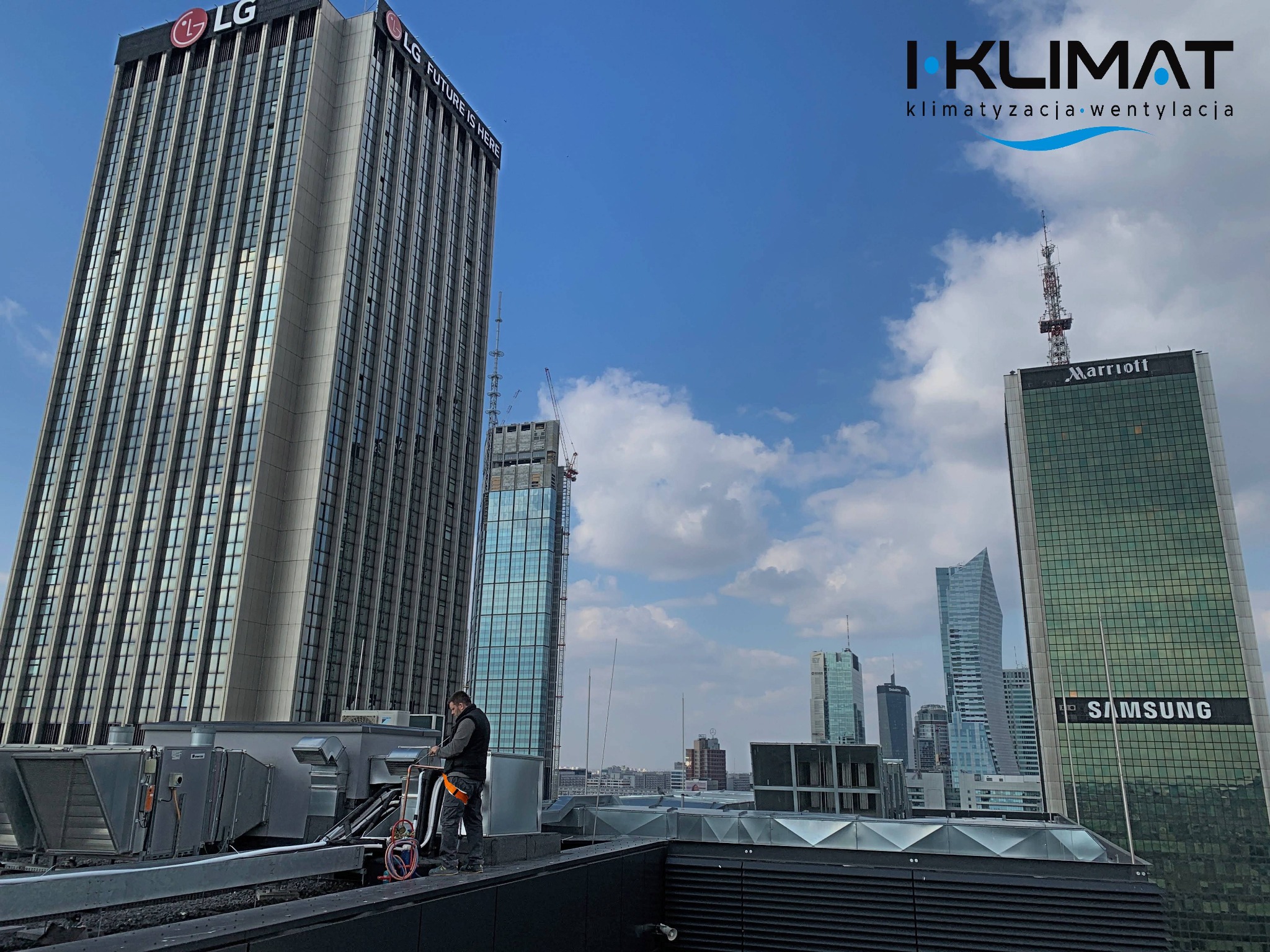 Pracownik serwisu w uprzęży na dachu budynku, wykonujący prace konserwacyjne przy systemie wentylacji i klimatyzacji na tle panoramy Warszawy z wieżowcami LG, Marriott i Samsung.