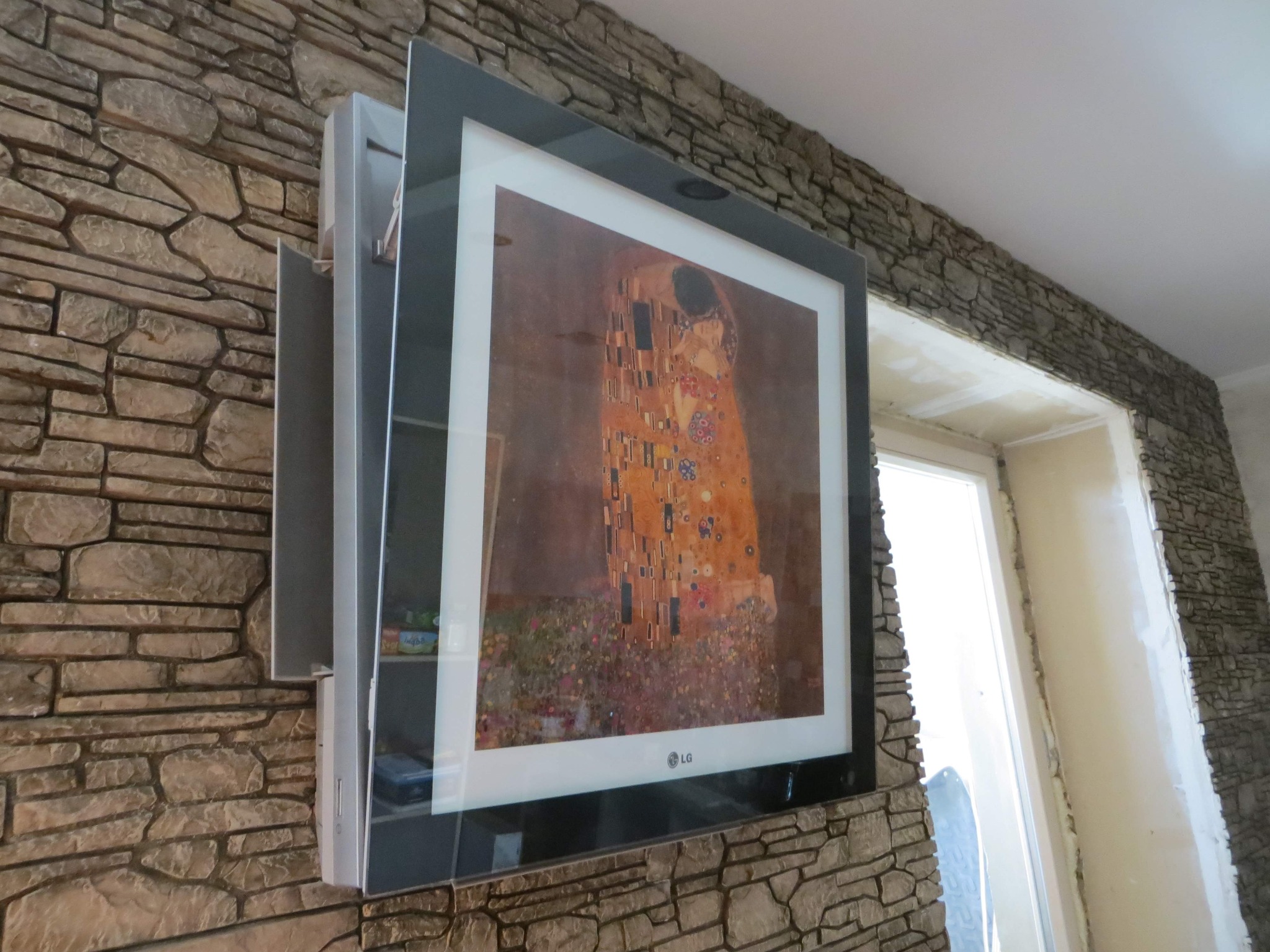 Telewizor z obrazem 'Pocałunek' Gustava Klimta, zamontowany na ścianie wyłożonej kamieniem dekoracyjnym, z widocznymi półkami po lewej stronie i oknem w tle.