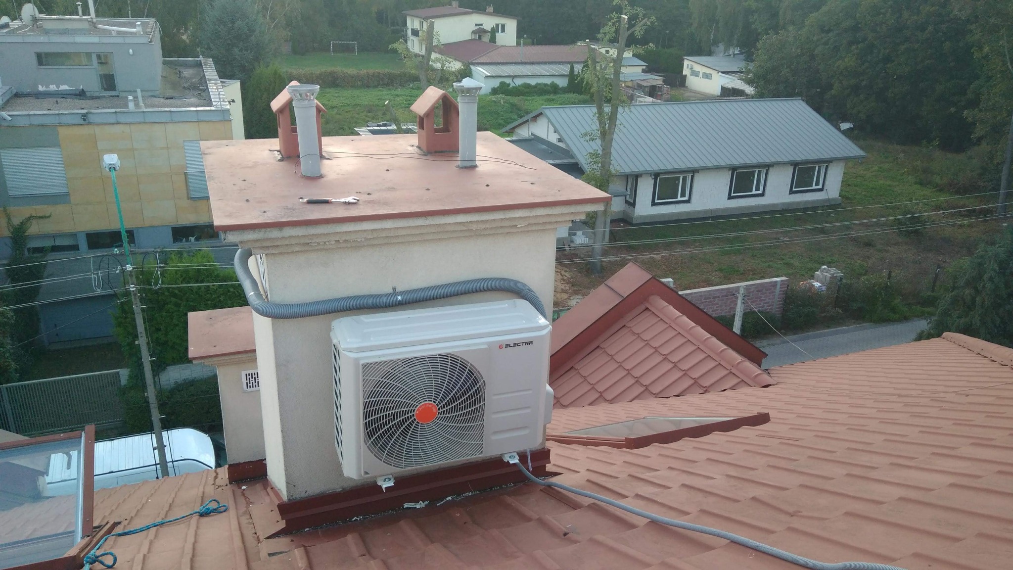 Biała jednostka zewnętrzna klimatyzatora marki Electra zamontowana na dachu budynku, widoczna szara rura odprowadzająca skropliny i fragment klucza francuskiego leżącego na dachu.