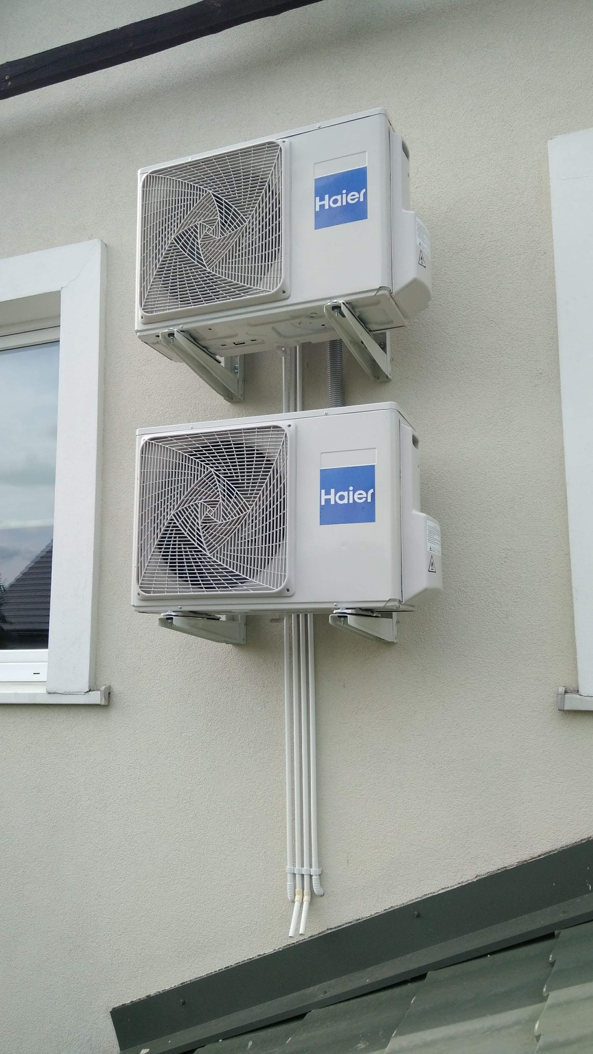 Dwie jednostki zewnętrzne klimatyzacji marki Haier zamontowane jedna nad drugą na elewacji budynku, widoczne rury instalacyjne i fragment okna.