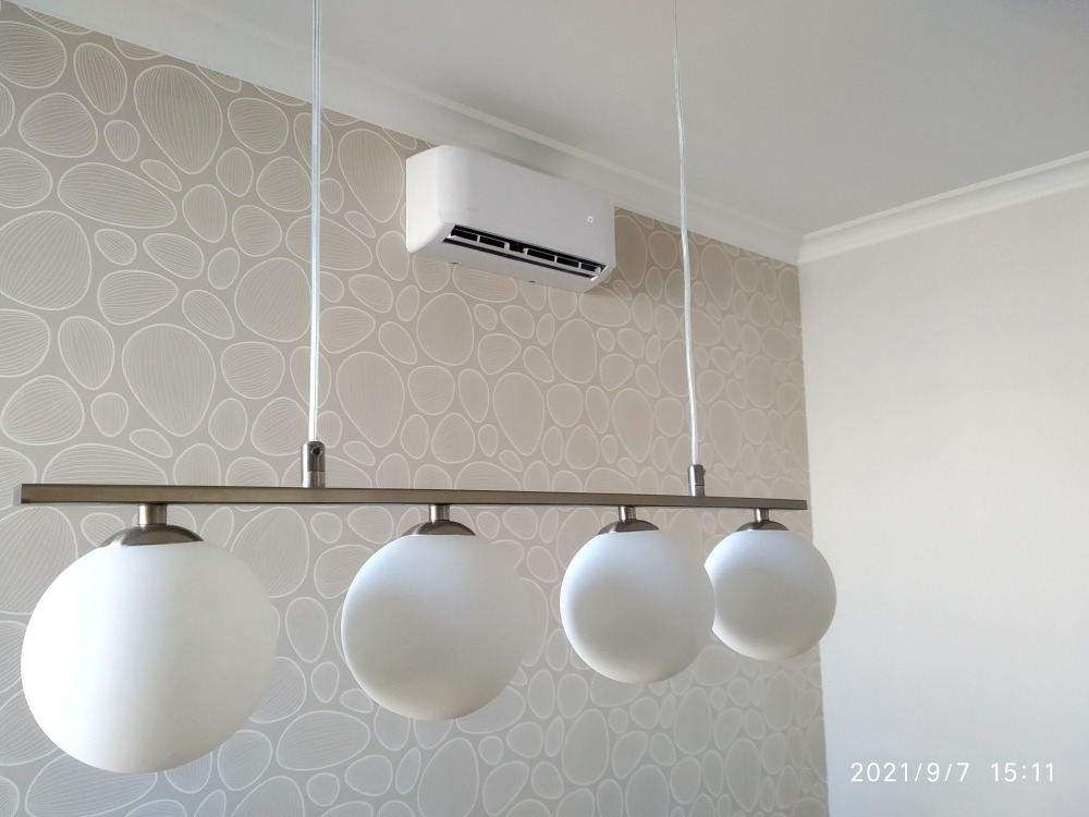 Biały klimatyzator ścienny zamontowany nad nowoczesną lampą z czterema mlecznymi kulami, na tle ściany w jasne, abstrakcyjne wzory.