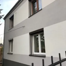 Dwupiętrowy budynek z elewacją w poziome pasy szarości i bieli, czarne rolety zewnętrzne, widok zza ogrodzenia.