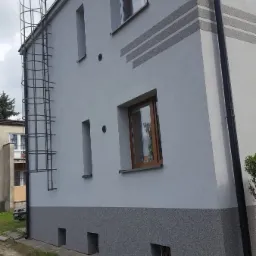 Szary budynek z ocieploną elewacją, widoczna metalowa drabina przeciwpożarowa, dekoracyjne pasy elewacyjne i cok&oacute;ł z fakturą baranka.