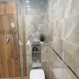 Nowoczesna łazienka z płytkami imitującymi marmur, kabina prysznicowa z drewnopodobnymi panelami, wisząca toaleta z mozaiką i roślina doniczkowa jako element dekoracyjny.