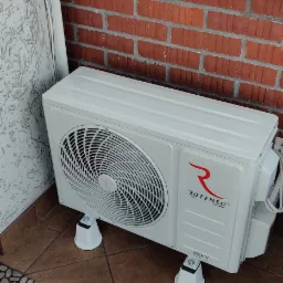 Biała jednostka zewnętrzna klimatyzacji Rotenso RONI Series zamontowana na balkonie z czerwoną cegłą, podniesiona na czterech białych wspornikach.