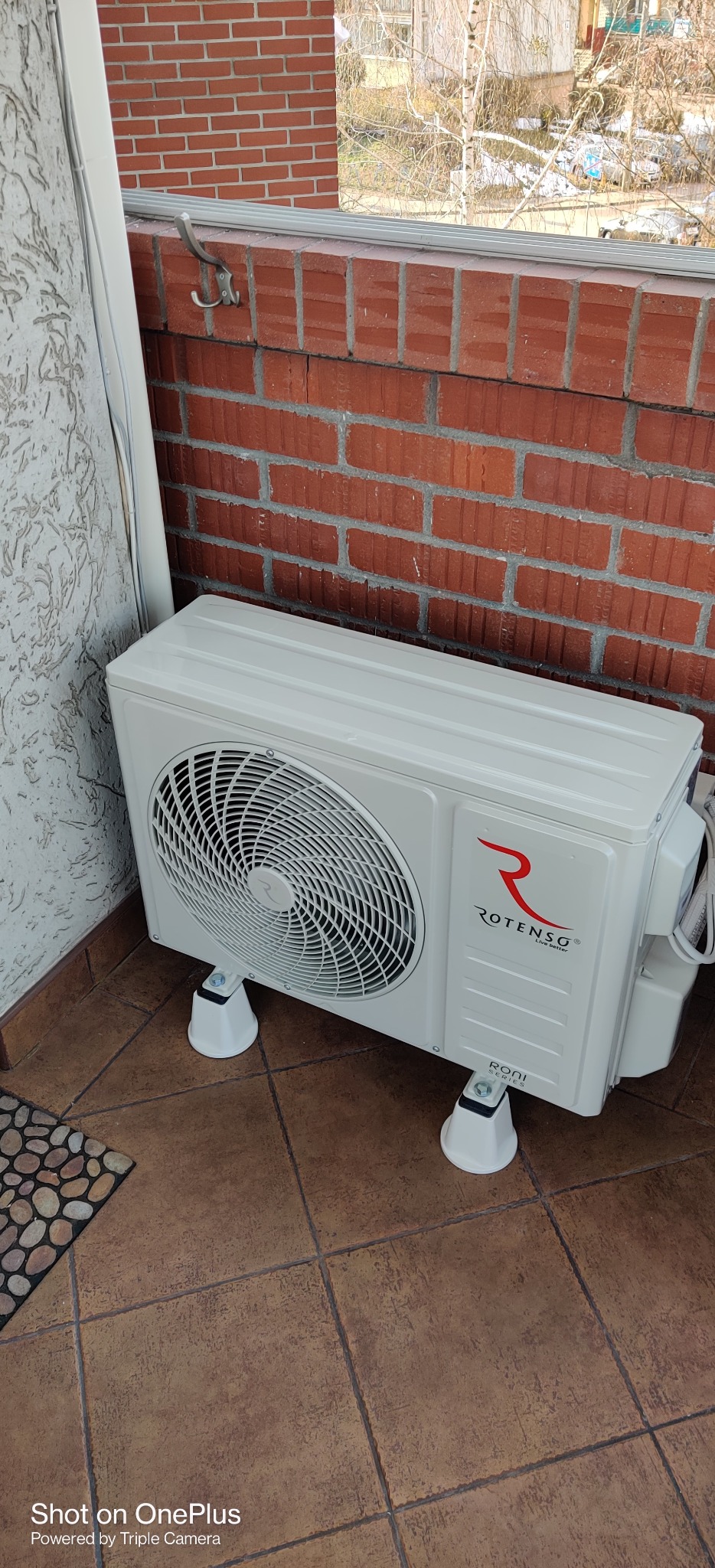 Biała jednostka zewnętrzna klimatyzacji Rotenso RONI Series zamontowana na balkonie z czerwoną cegłą, podniesiona na czterech białych wspornikach.