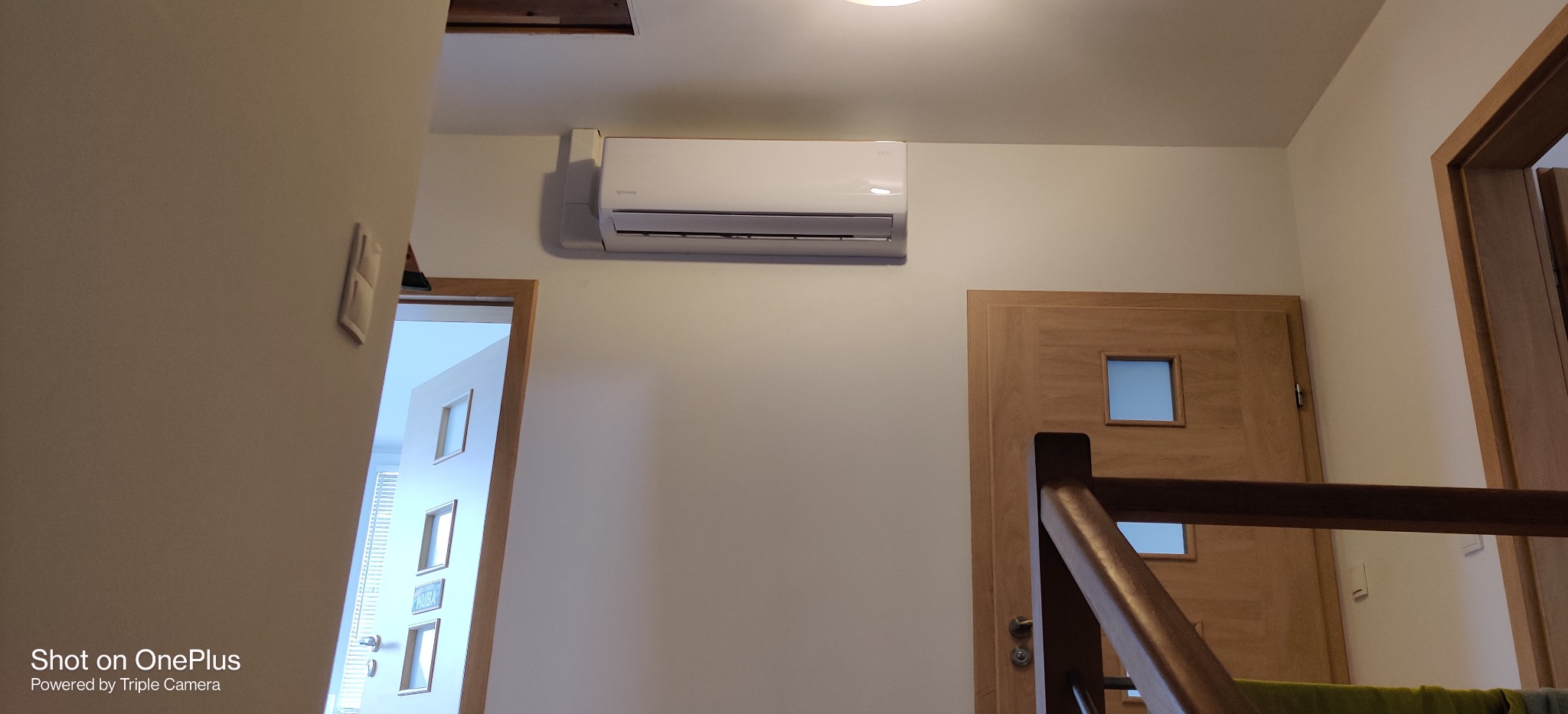 Biała klimatyzacja ścienna zamontowana w jasnym wnętrzu domu, widoczna obok drewnianych drzwi i fragmentu schodów.