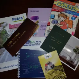 Stos różnorodnych materiałów drukowanych, w tym broszury, czasopismo z krzyżówkami, zaproszenie, cennik produktów i katalogi, ułożone na ciemnym tle.