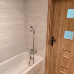 Solid-tech - Wykończona łazienka z wanną, chromowaną baterią i słuchawką prysznicową na tle jasnych, marmurkowych płytek ściennych oraz drewnianych drzwi z szybkami.