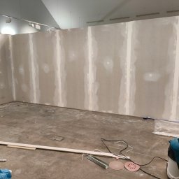 Solid-tech - Wnętrze w trakcie prac wykończeniowych: ściany z płyt kartonowo-gipsowych z widocznymi łączeniami przed szpachlowaniem, na podłodze narzędzia, listwy przypodłogowe i odkurzacz przemysłowy Makita.