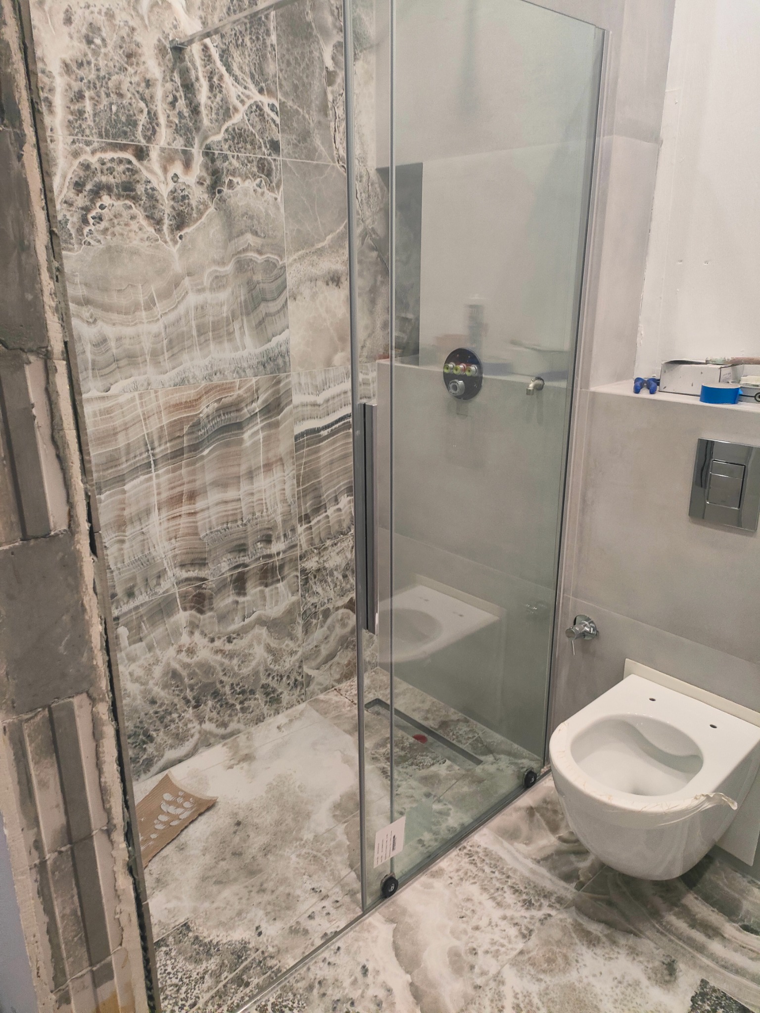 Montaż kabiny prysznicowej z drzwiami szklanymi i wykończeniem ściany imitującym marmur, widoczny fragment niezabezpieczonej ściany oraz toaleta w folii ochronnej, w trakcie remontu łazienki.