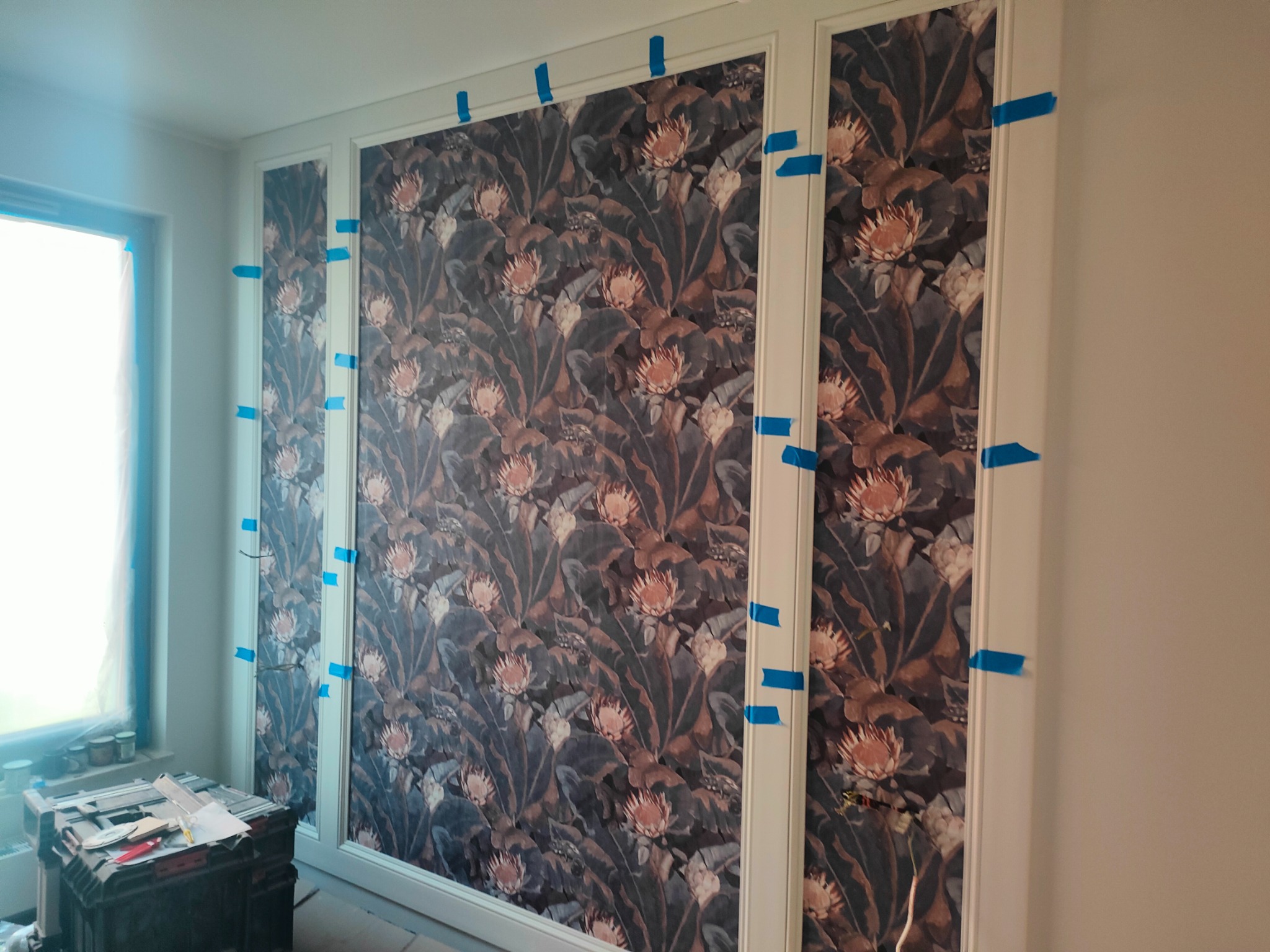 Montaż dekoracyjnych paneli ściennych z motywem kwiatowym, zabezpieczonych taśmą malarską na białych listwach, widoczna skrzynka narzędziowa i okno w tle.