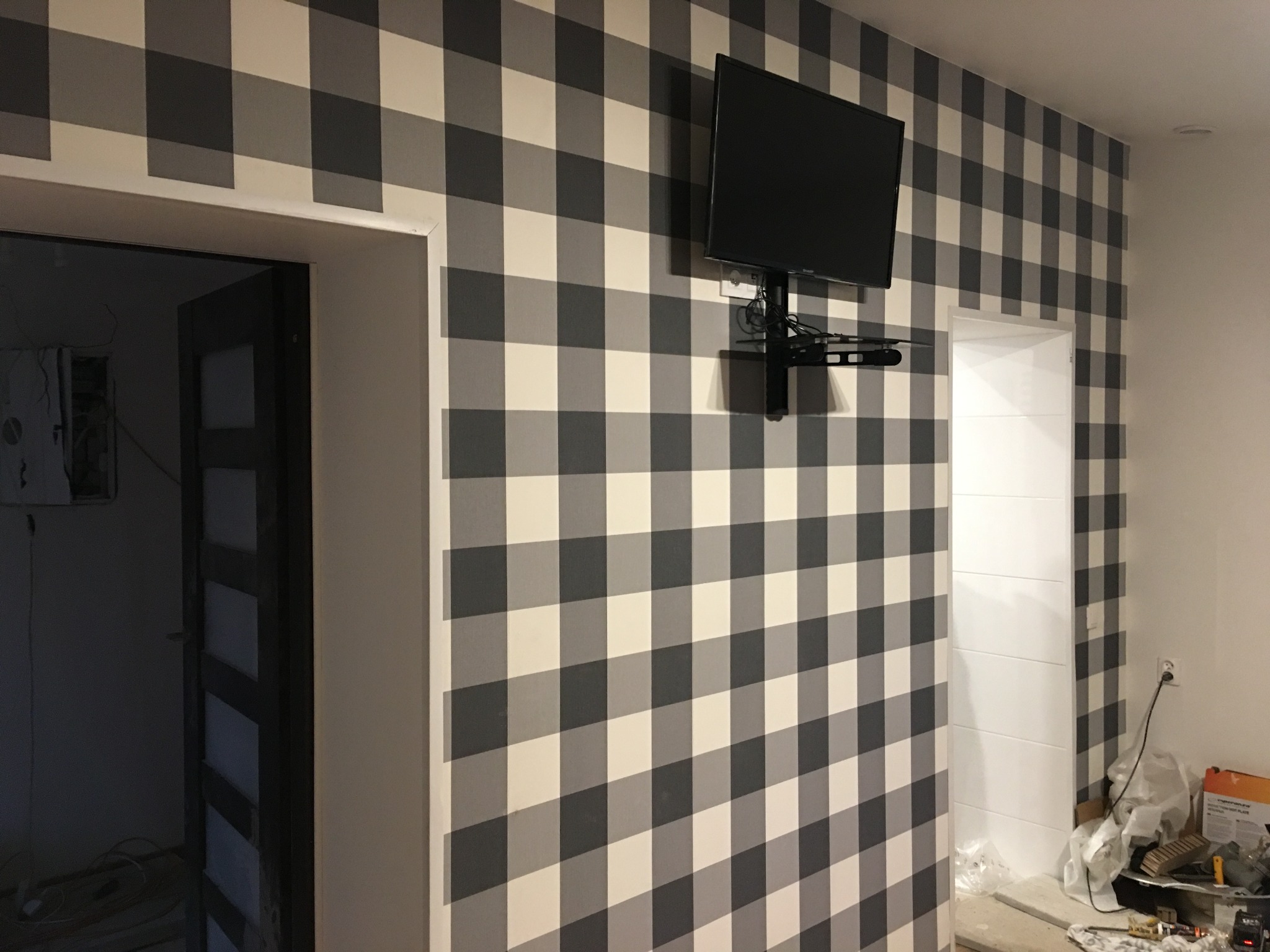Ściana w szarą kratę z zamontowanym telewizorem na uchwycie, widoczne fragmenty ościeżnic drzwiowych i drobne prace wykończeniowe w pomieszczeniu.