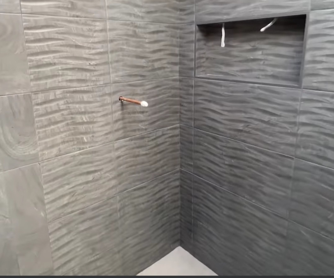 Szare, faliste płytki ceramiczne w rogu łazienki, z wnęką w ścianie, widoczny fragment rury wystającej ze ściany.