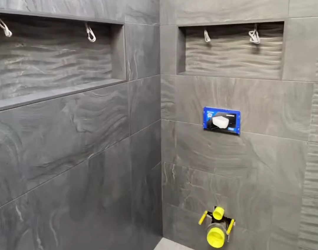 Szare płytki imitujące kamień z wnękami i widoczną instalacją podtynkową WC w trakcie montażu.