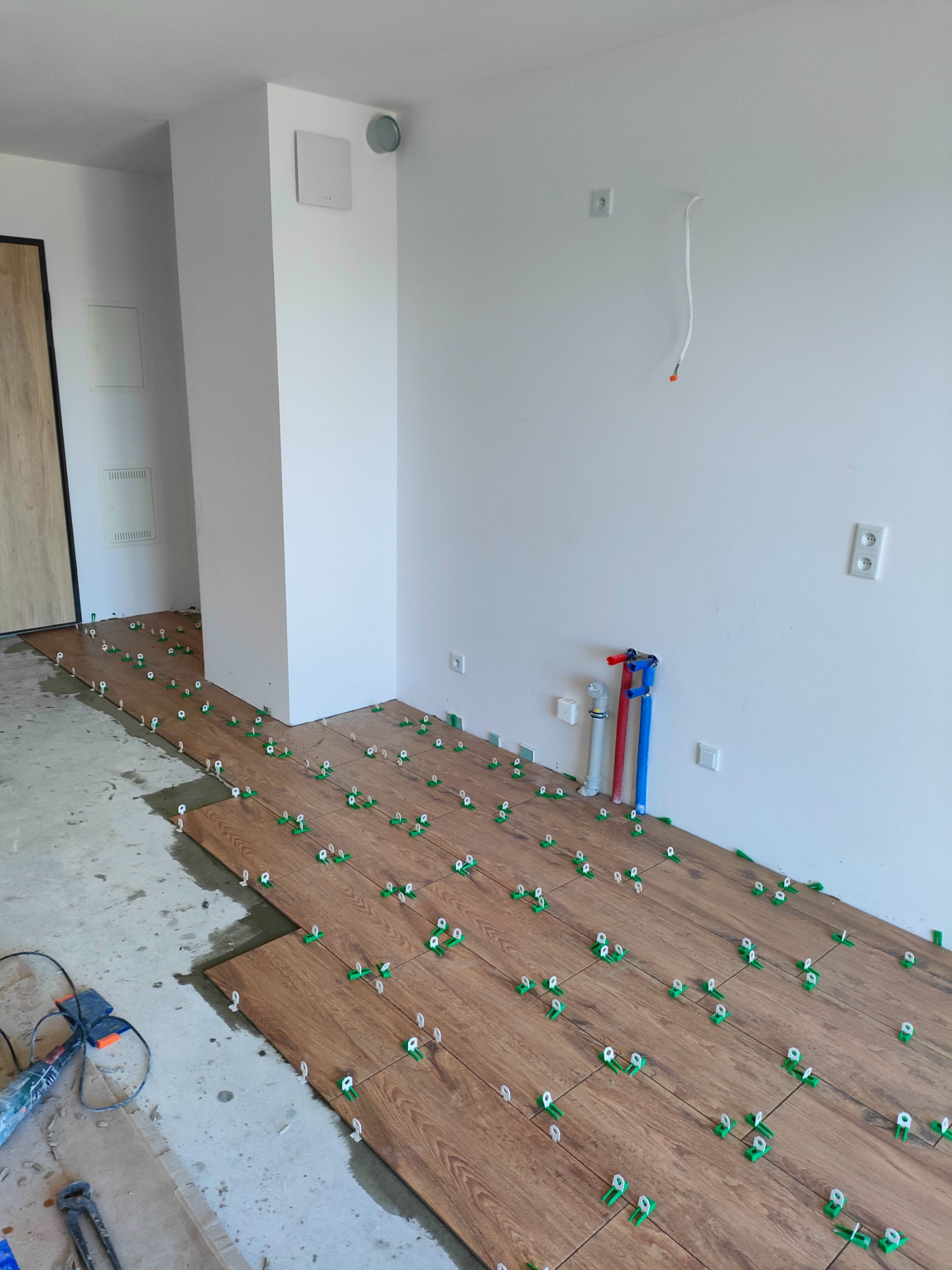 Układanie płytek podłogowych imitujących drewno, z użyciem systemu poziomowania płytek (zielone klipsy i białe kliny), widoczne fragmenty nieukładanych płytek oraz narzędzia pracy.