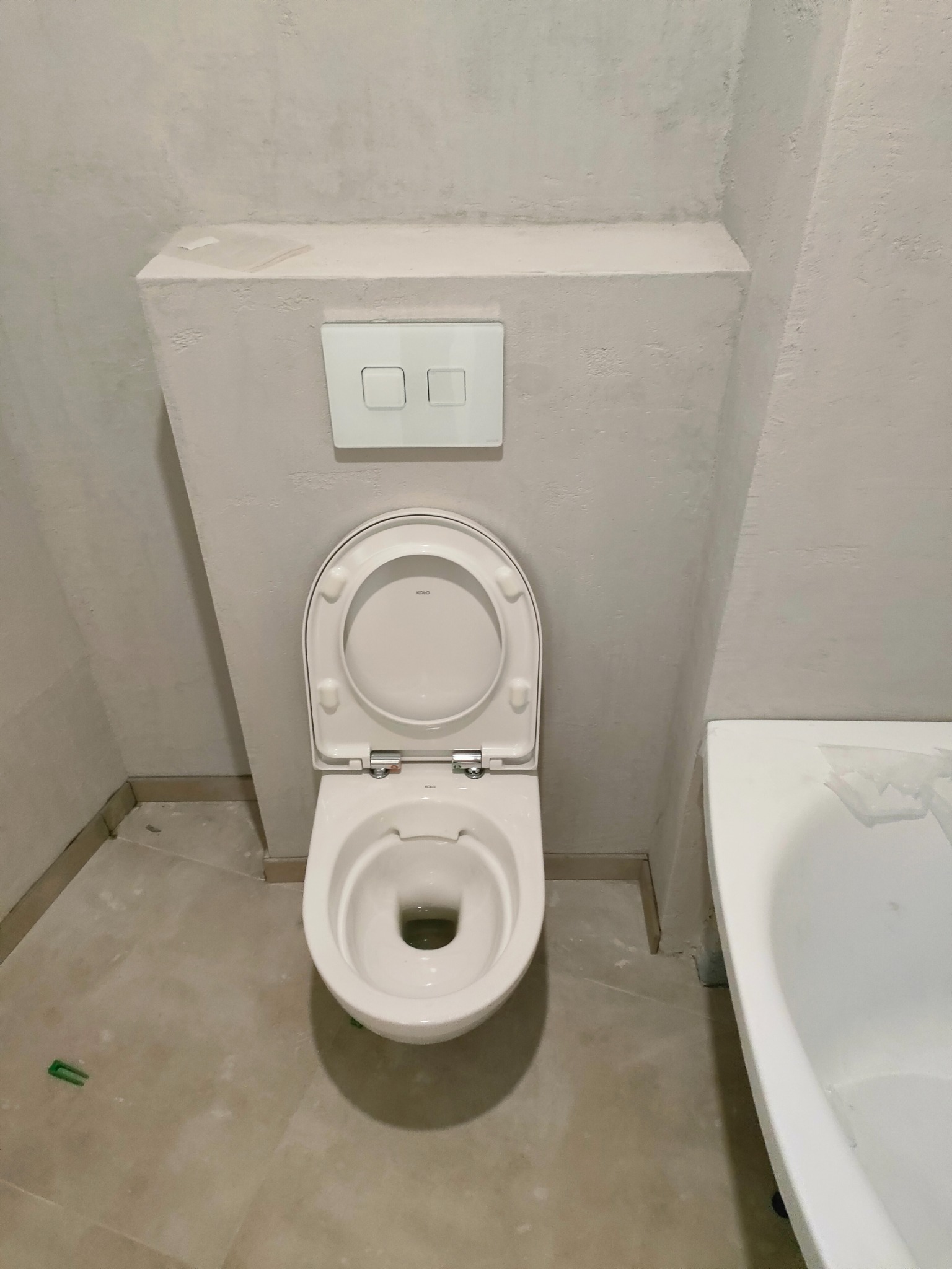 Biała toaleta z otwartą klapą w surowym wnętrzu łazienki, widoczna zabudowa geberitu z przyciskami spłukiwania i fragment wanny, ściany w trakcie wykończenia.