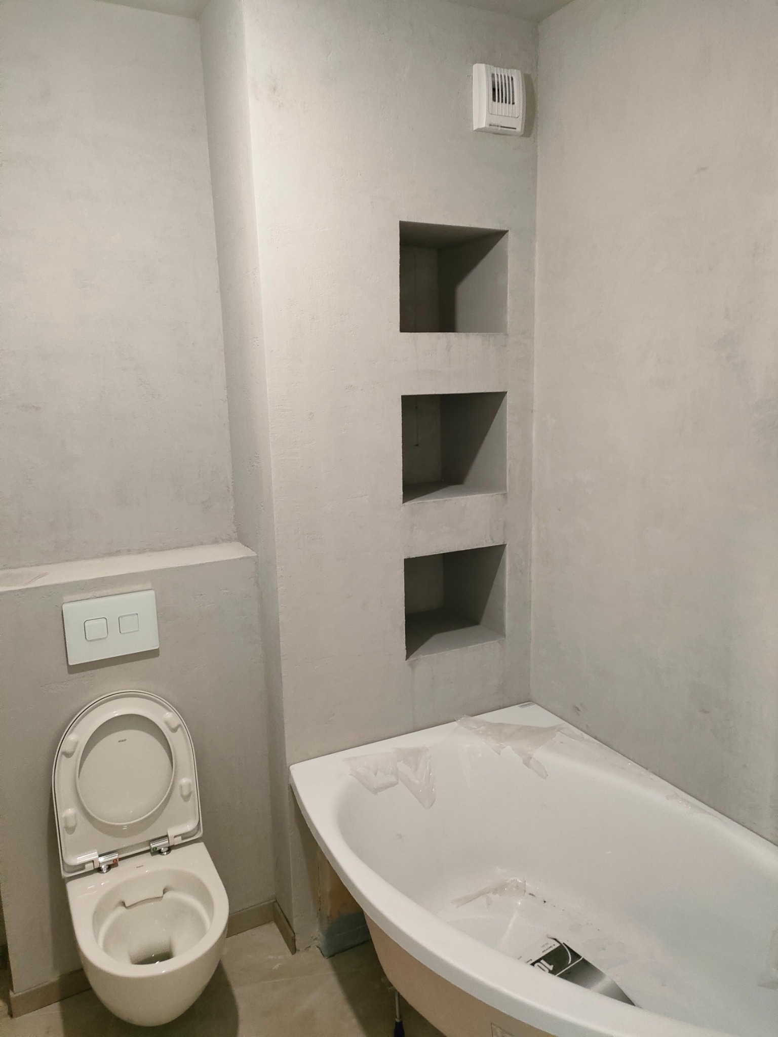 Łazienka w trakcie wykończenia w Gdańsku: widoczne gładzie na ścianach z wnękami, częściowo zabezpieczona wanna i wisząca toaleta z otwartą klapą.