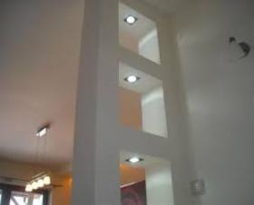 Biała ściana z trzema wnękami oświetlonymi punktowymi lampami sufitowymi, widok z dołu, w tle fragment salonu z lampą wiszącą.