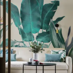 Salon w Warszawie z kremową sofą, okrągłym czarnym stolikiem kawowym, poduszkami w odcieniach zieleni i tapetą z motywem tropikalnych liści.