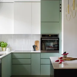 Nowoczesna kuchnia z frontami w kolorze pistacjowym i białym, płytki w pionowy wzór, czarny ekspres do kawy na blacie, złote wiszące lampy, czerwone papryczki chili jako dekoracja.