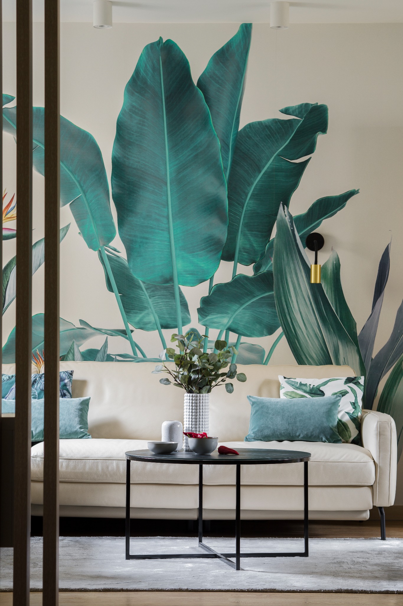 Salon w Warszawie z kremową sofą, okrągłym czarnym stolikiem kawowym, poduszkami w odcieniach zieleni i tapetą z motywem tropikalnych liści.