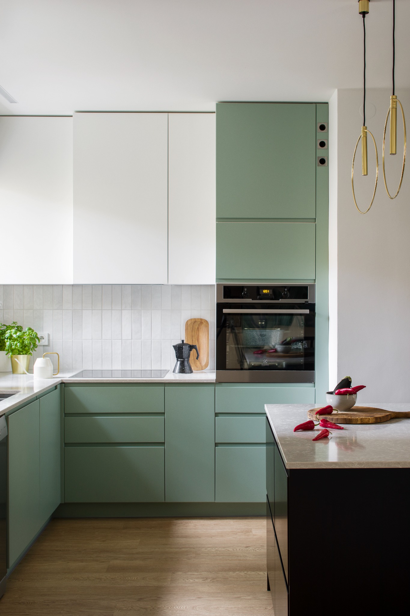 Nowoczesna kuchnia z frontami w kolorze pistacjowym i białym, płytki w pionowy wzór, czarny ekspres do kawy na blacie, złote wiszące lampy, czerwone papryczki chili jako dekoracja.